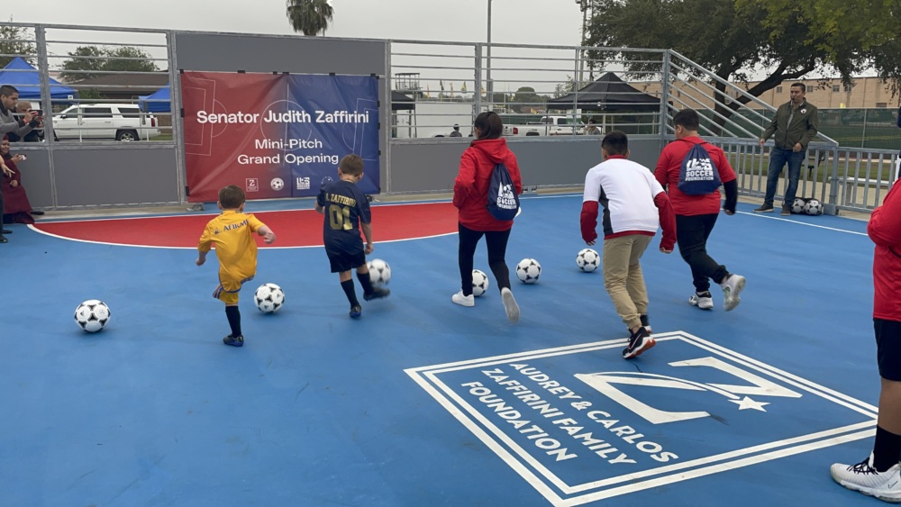 Five kids kick soccer balls into a goal that is covered by a banner to officially open a new mini-pitch.